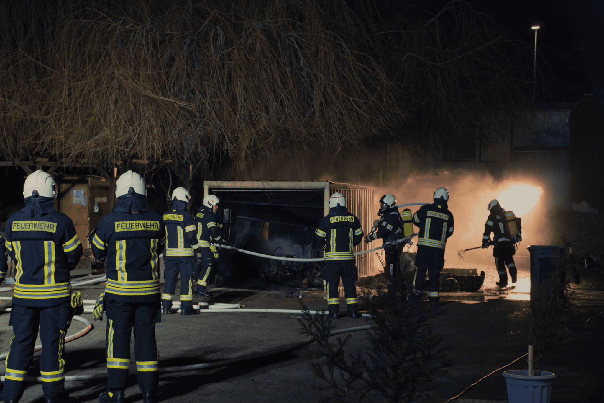 Feuerwehrleute bekämpfen einen nächtlichen Garagenbrand mit Schläuchen und Werkzeugen.