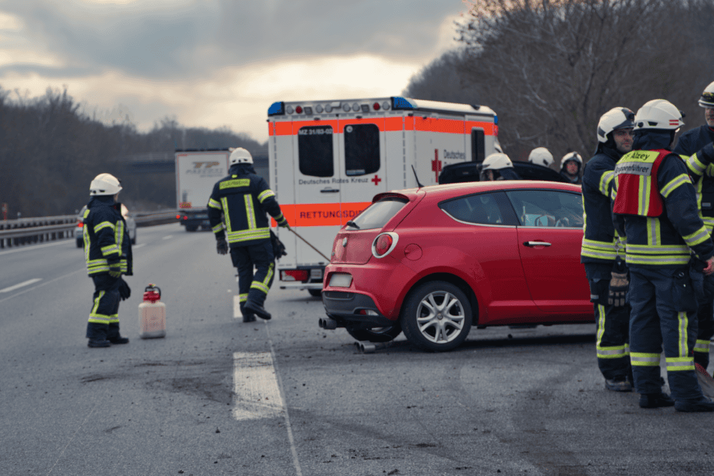 Feuerwehrleute sichern die Unfallstelle auf einer Autobahn neben einem roten PKW, ein Rettungswagen steht im Hintergrund.