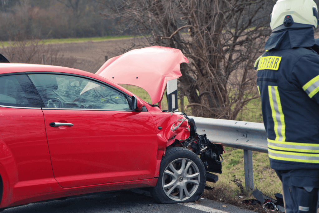 Ein roter PKW mit beschädigter Front steht an einer Leitplanke, ein Feuerwehrmann steht daneben.