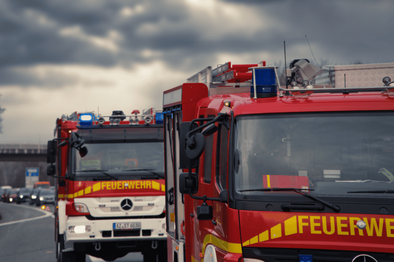 Zwei rote Feuerwehrfahrzeuge mit eingeschaltetem Blaulicht auf einer Autobahn unter einem wolkigen Himmel.