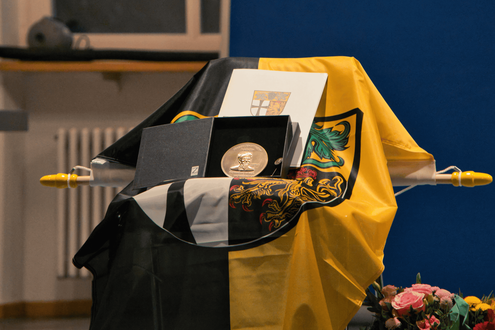 Eine goldene Medaille in einer Box, zusammen mit einer Urkunde auf einer schwarz-gelben Flagge drapiert.