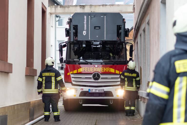 Großes Hubrettungsfahrzeug fährt durch eine schmale Gasse, während mehrere Feuerwehrleute den Weg sichern.